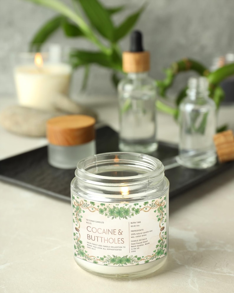 Op de afbeelding: Een brandende glazen potkaars met een label met de tekst "Cocaine & Buttholes" omgeven door een groen bloemmotief. De kaars maakt deel uit van de Veteran Candles-collectie. Andere glazen flessen en een pot met houten deksel staan op de achtergrond.