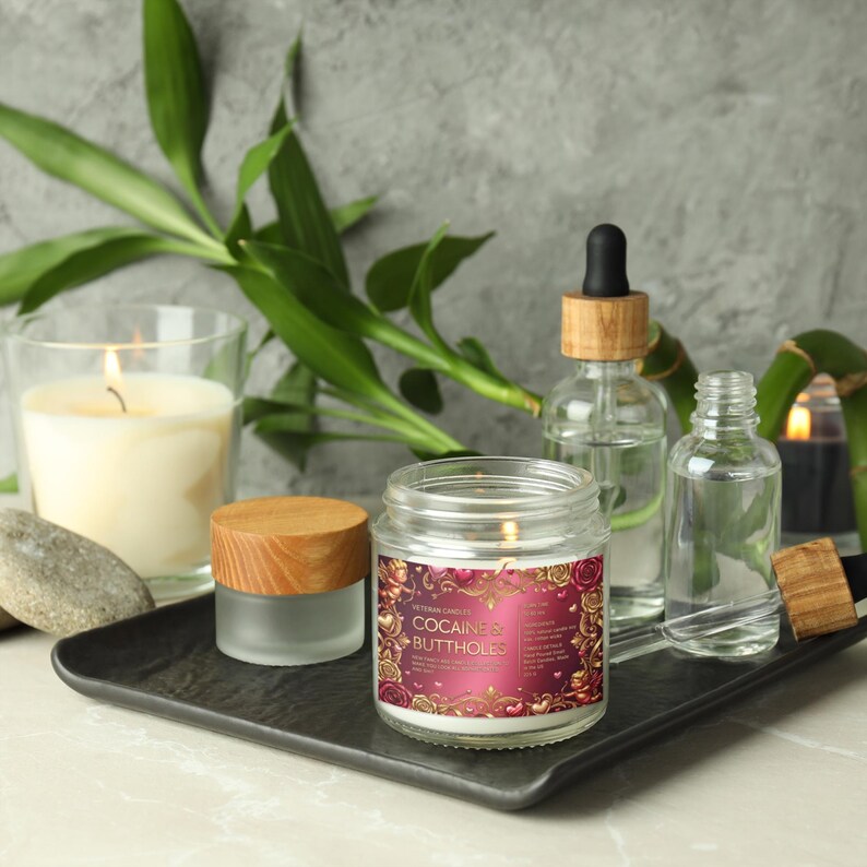Peut inclure: Une bougie allum&eacute;e dans un bocal en verre avec une &eacute;tiquette indiquant "COCAINE & BUTTHOLES" est pos&eacute;e sur un plateau noir, &agrave; c&ocirc;t&eacute; d'autres bougies, de flacons en verre et de bambou. La bougie a un couvercle en bois et est entour&eacute;e d'autres objets d&eacute;coratifs.