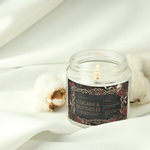 May include: A lit, clear glass jar candle with a black label that reads "COCAINE & BUTTHOLES" in white text. The label has a decorative border with roses and skulls. The candle sits on a white surface with cotton buds.