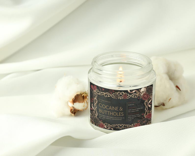 May include: A lit, clear glass jar candle with a black label that reads "COCAINE & BUTTHOLES" in white text. The label has a decorative border with roses and skulls. The candle sits on a white surface with cotton buds.