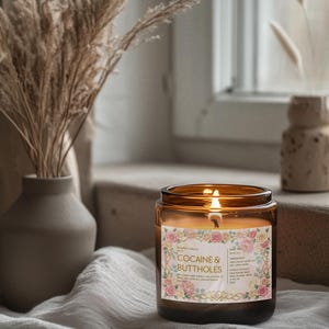 Pu&ograve; includere: Una candela accesa in vetro ambrato con un'etichetta floreale che recita "Cocaine & Buttholes". La candela &egrave; su un panno bianco vicino a una finestra. Un vaso con piante secche &egrave; sullo sfondo.