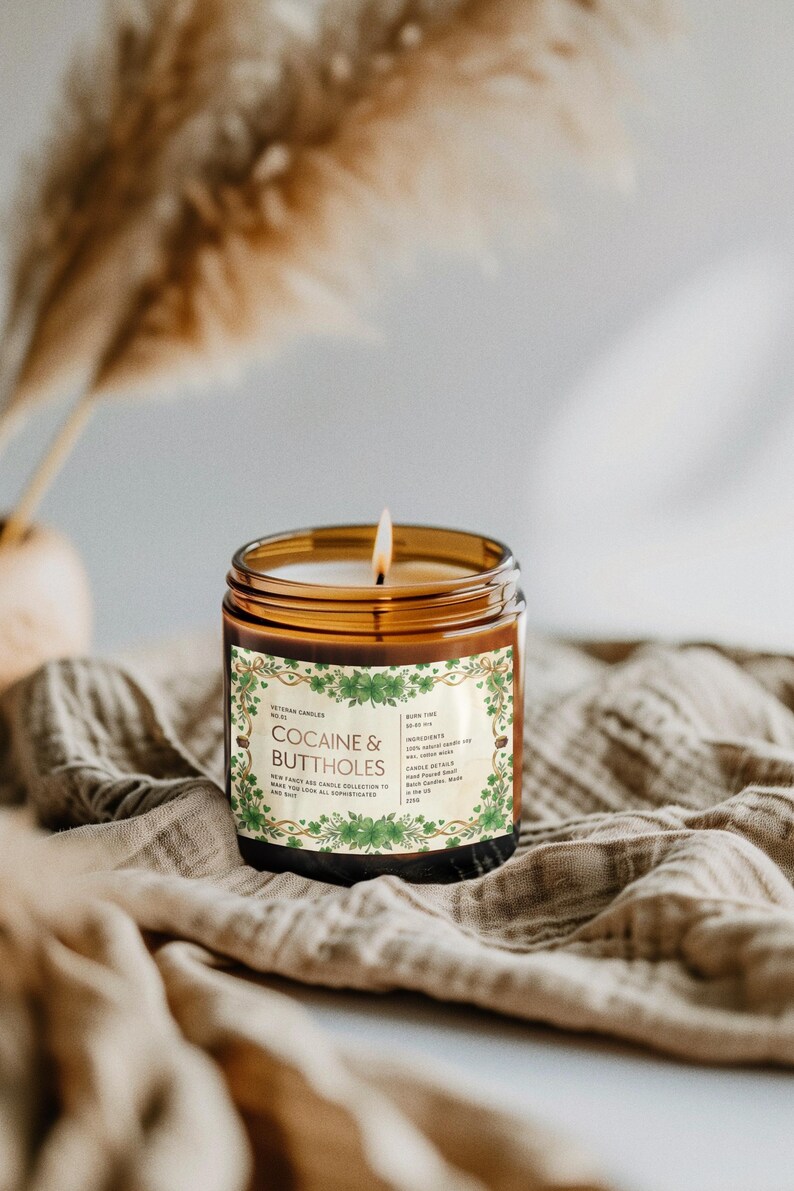 Może przedstawiać: Zapalone świeca w brązowym szklanym słoiku z etykietą z napisem "Cocaine & Buttholes". Etykieta ma zielony kwiatowy wz&oacute;r. Świeca stoi na beżowej tkaninie.
