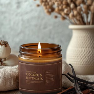 May include: A lit brown glass candle with the text "Cocaine & Buttholes" on a brown label. The candle sits on a wooden surface with cinnamon sticks and vanilla beans. A vase with dried flowers is in the background.