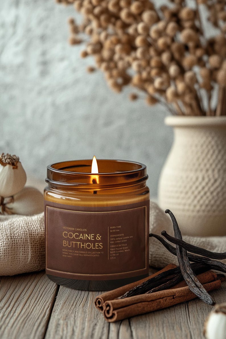 May include: A lit brown glass candle with the text "Cocaine & Buttholes" on a brown label. The candle sits on a wooden surface with cinnamon sticks and vanilla beans. A vase with dried flowers is in the background.