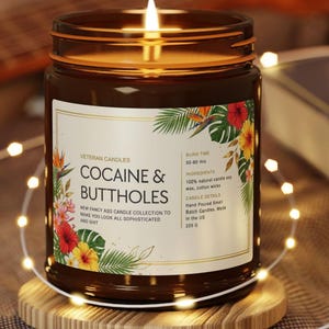 Może przedstawiać: Zapalone brązowe szklane naczynie na świece z etykietą z napisem "COCAINE & BUTTHOLES". Etykieta jest ozdobiona tropikalnymi kwiatami i liśćmi. Świeca stoi na drewnianej podstawce i jest otoczona łańcuchem świetlnym. Świeca jest częścią "NEW FANCY ASS CANDLE COLLECTION."