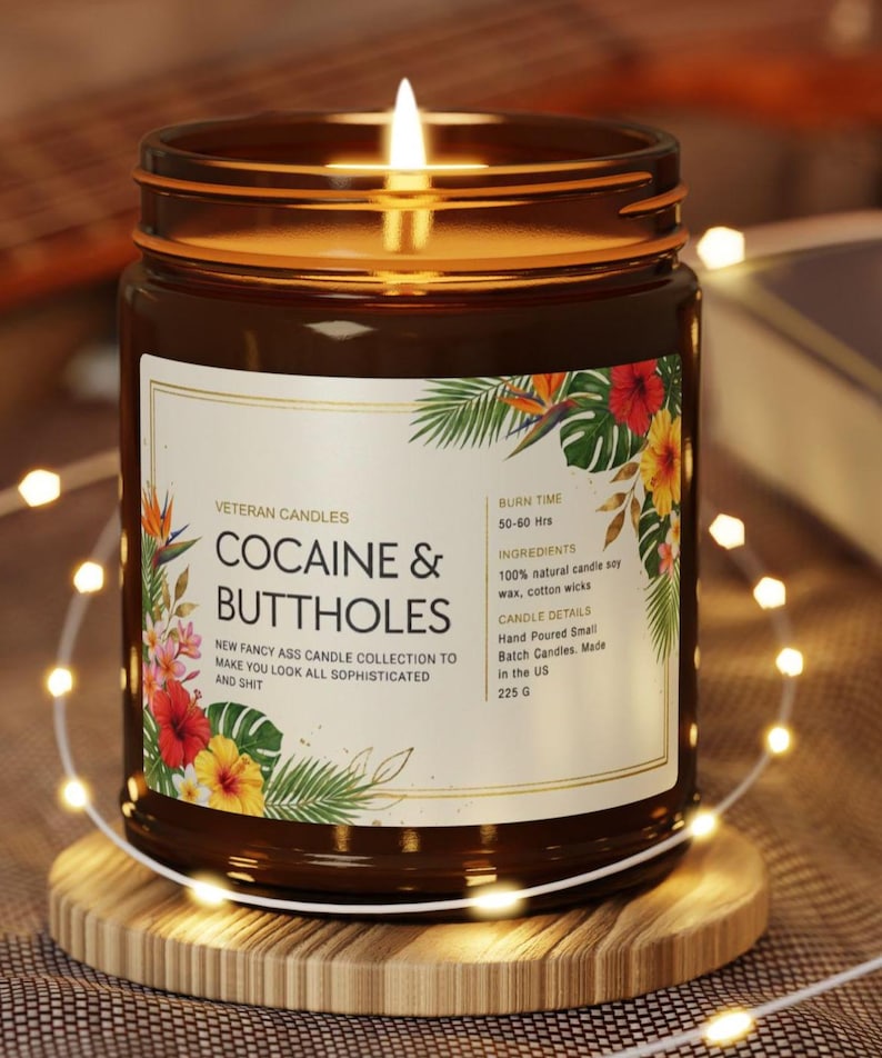 Może przedstawiać: Zapalone brązowe szklane naczynie na świece z etykietą z napisem "COCAINE & BUTTHOLES". Etykieta jest ozdobiona tropikalnymi kwiatami i liśćmi. Świeca stoi na drewnianej podstawce i jest otoczona łańcuchem świetlnym. Świeca jest częścią "NEW FANCY ASS CANDLE COLLECTION."