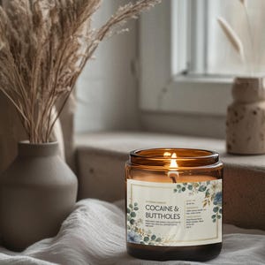 May include: A lit amber glass candle with the text "COCAINE & BUTTHOLES" on the label, surrounded by floral designs. The candle sits on a draped surface near a window, with a vase of dried plants in the background.