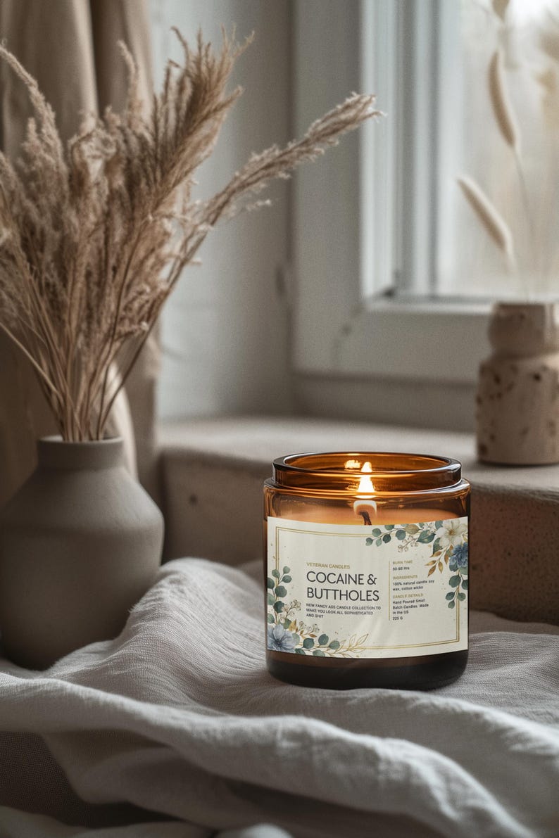 May include: A lit amber glass candle with the text "COCAINE & BUTTHOLES" on the label, surrounded by floral designs. The candle sits on a draped surface near a window, with a vase of dried plants in the background.