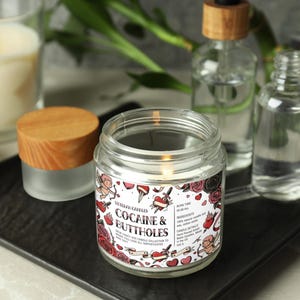 May include: A lit glass jar candle with a label that reads "Cocaine & Buttholes" surrounded by tattoo-style roses and hearts. Other items include a small jar with a wooden lid, two clear bottles, and a lit candle in a glass.