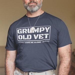 Könnte beinhalten: Marineblaues T-Shirt mit dem Text "GRUMPY OLD VET" in einem weißen Rechteck. Darunter steht "LEAVE ME ALONE". Die Person trägt eine Halskette mit Erkennungsmarke und eine Uhr.