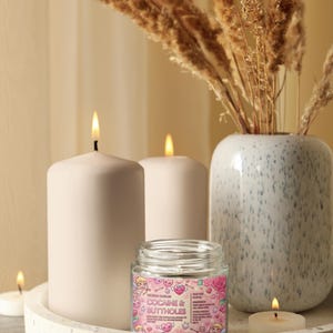 May include: A decorative arrangement featuring lit candles, a glass jar with a pink label, and a vase with dried plants. The jar's label reads "COCAINE & BUTTHOLES" with cartoon graphics. The candles are white and cylindrical, and the vase is speckled.