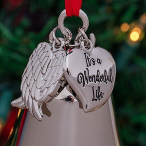 May include: Silver Christmas ornament with a heart and angel wing design. The heart-shaped ornament has the phrase "It's a Wonderful Life" in black script. A red ribbon is attached for hanging.