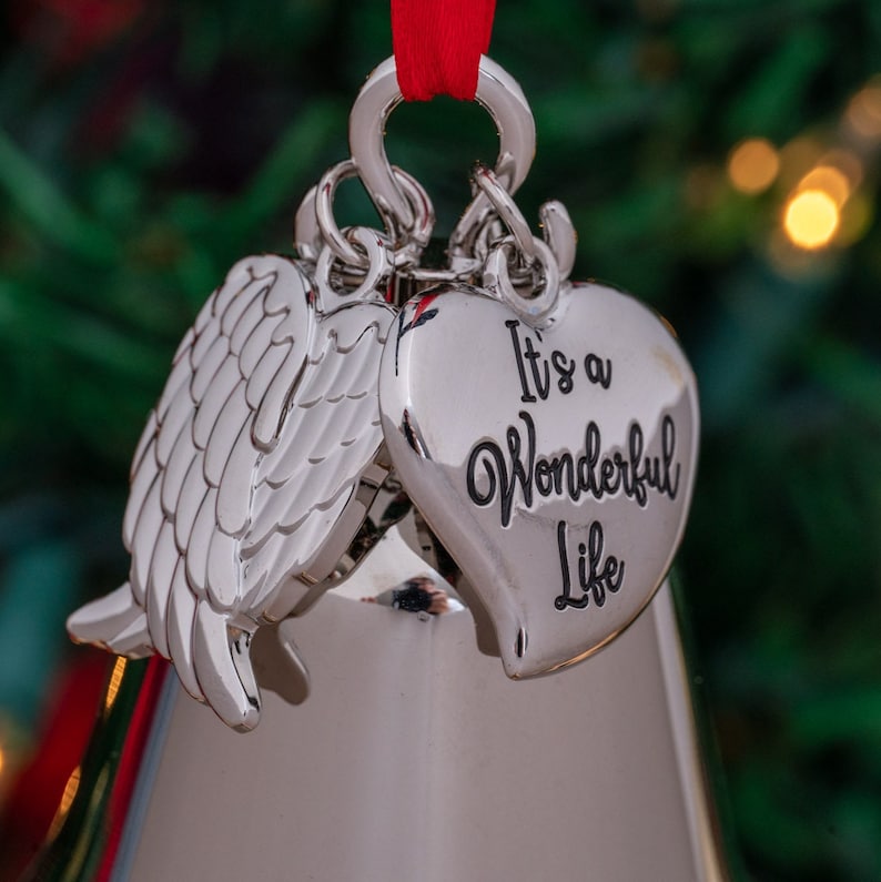 May include: Silver Christmas ornament with a heart and angel wing design. The heart-shaped ornament has the phrase "It's a Wonderful Life" in black script. A red ribbon is attached for hanging.