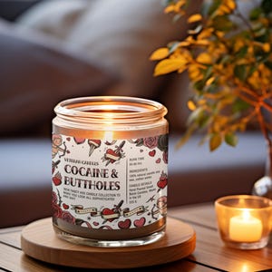 May include: A glass jar candle with the text "COCAINE & BUTTHOLES" and other text. The candle has a burn time of 50-60 hours and is made with natural soy wax and cotton wicks. The candle is on a wooden coaster with a lit votive candle nearby.