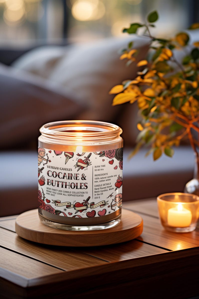 May include: A glass jar candle with the text "COCAINE & BUTTHOLES" and other text. The candle has a burn time of 50-60 hours and is made with natural soy wax and cotton wicks. The candle is on a wooden coaster with a lit votive candle nearby.