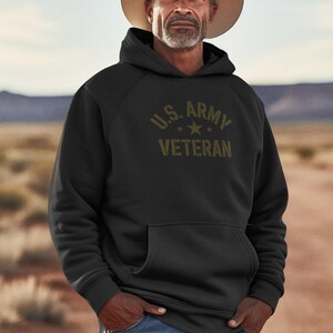 Puede incluir: Sudadera negra con el texto "U.S. ARMY VETERAN" en verde oliva. La sudadera tiene un bolsillo delantero y una capucha con cord&oacute;n. La persona de la imagen lleva un sombrero de vaquero y vaqueros azules.