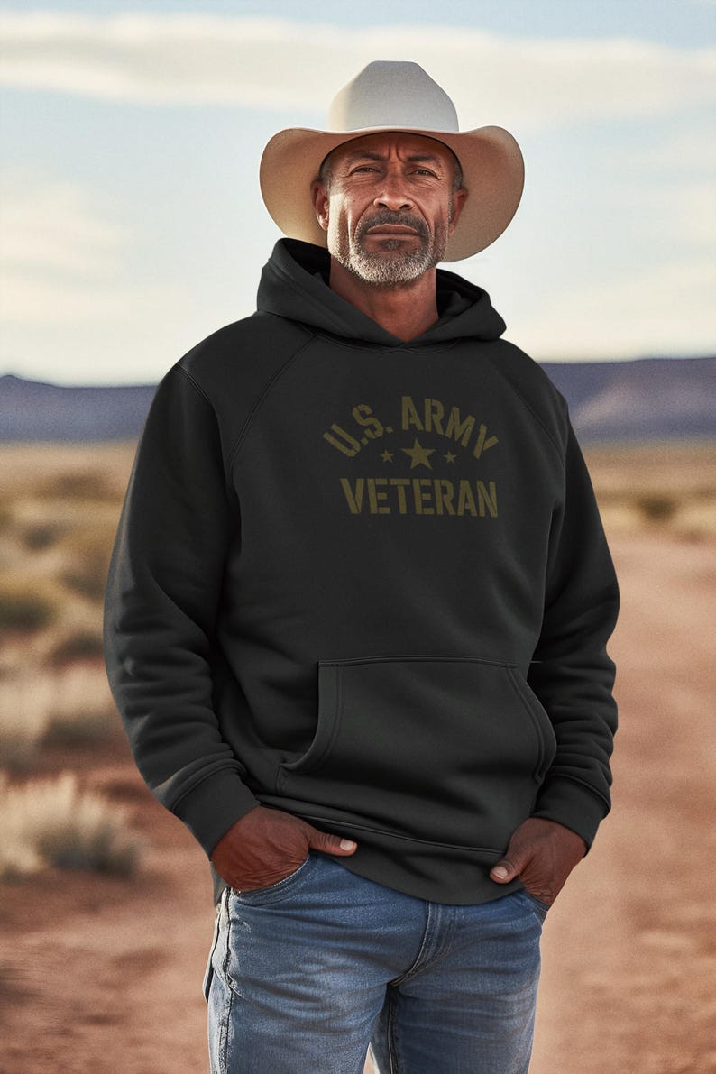 Puede incluir: Sudadera negra con el texto "U.S. ARMY VETERAN" en verde oliva. La sudadera tiene un bolsillo delantero y una capucha con cord&oacute;n. La persona de la imagen lleva un sombrero de vaquero y vaqueros azules.