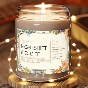 May include: A lit, grey-brown candle in a glass jar with a label that reads "NIGHTSHIFT & C. DIFF." The label has floral accents and text that says "New Fancy Ass Nurse Candle Collection." The candle sits on a wooden coaster.