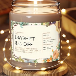 May include: A lit, brown glass jar candle with a floral label. The label reads "DAYSHIFT & C. DIFF" and "NEW FANCY ASS NURSE CANDLE COLLECTION TO MAKE YOU LOOK ALL SOPHISTICATED AND SHIT." The candle sits on a wooden coaster.