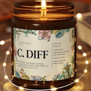May include: A lit brown glass jar candle with a label that reads "C. DIFF" and "New Fancy Ass Nurse Candle Collection". The label has floral and medical-themed illustrations. The candle sits on a wooden coaster, surrounded by fairy lights.