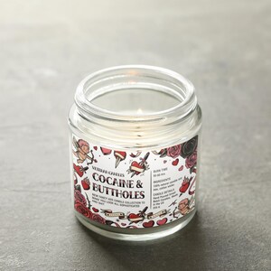 Peut inclure: Une bougie en pot de verre transparent allum&eacute;e avec une &eacute;tiquette qui dit "Cocaine & Buttholes". L'&eacute;tiquette pr&eacute;sente des illustrations de style tatouage de c&oelig;urs, de roses et de ch&eacute;rubins. La bougie fait partie de la "New Fancy Ass Candle Collection."