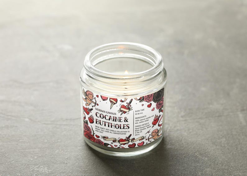 Peut inclure: Une bougie en pot de verre transparent allum&eacute;e avec une &eacute;tiquette qui dit "Cocaine & Buttholes". L'&eacute;tiquette pr&eacute;sente des illustrations de style tatouage de c&oelig;urs, de roses et de ch&eacute;rubins. La bougie fait partie de la "New Fancy Ass Candle Collection."