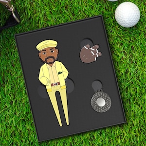 May include: A black box set on green grass containing golf accessories. The set includes a yellow cartoon figure, a brown hand, and a silver divot tool. Two white golf balls are visible in the background. The cartoon figure is wearing a yellow outfit.