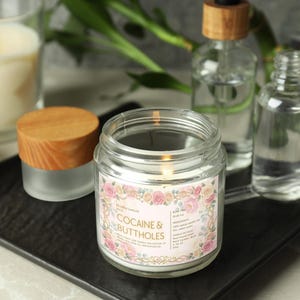 Puede incluir: Una vela encendida en un tarro de cristal transparente con tapa de madera, etiquetada "COCAINE & BUTTHOLES" con detalles florales. Otras botellas de vidrio y una vela est&aacute;n en el fondo, todo sobre una bandeja negra.