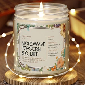 May include: A lit glass jar candle with a label that reads "Microwave Popcorn & C. Diff." The label includes floral designs and text indicating a 45-50 hour burn time. The candle sits on a wooden coaster, surrounded by string lights.