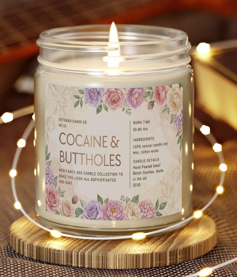 May include: A lit glass jar candle with a floral label that reads "COCAINE & BUTTHOLES." The candle is on a wooden coaster and surrounded by string lights. The label also includes the text "NEW FANCY ASS CANDLE COLLECTION TO MAKE YOU LOOK ALL SOPHISTICATED AND SHIT."