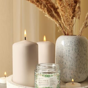 Może przedstawiać: Dekoracyjna aranżacja z zapalonymi świecami i małą świecą w słoiku z napisem "COCAINE & BUTTHOLES". Świece znajdują się na tacy z lastryko, z wazonem z suszonymi roślinami w tle. Scena oświetlona jest ciepłym, miękkim światłem.