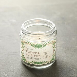 Może przedstawiać: Zapalone, przezroczyste szklane świece w słoiku z etykietą z napisem "Cocaine & Buttholes". Etykieta ma zielony kwiatowy wz&oacute;r. Świeca jest od Veteran Candles. Świeca stoi na szarej powierzchni.