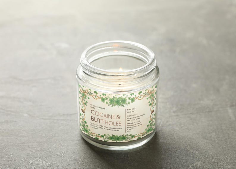 Może przedstawiać: Zapalone, przezroczyste szklane świece w słoiku z etykietą z napisem "Cocaine & Buttholes". Etykieta ma zielony kwiatowy wz&oacute;r. Świeca jest od Veteran Candles. Świeca stoi na szarej powierzchni.