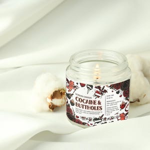 Może przedstawiać: Świeca w przezroczystym szklanym słoiku z zapalonym knotem. Etykieta głosi "Cocaine & Buttholes" z wzorem w stylu tatuażu. Świeca stoi na białym tle z kuleczkami bawełny.