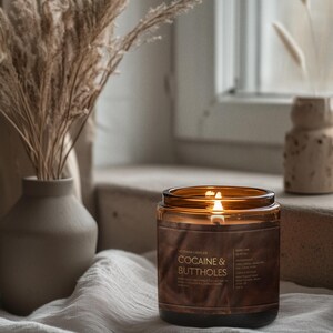 May include: A lit amber glass candle jar with the text "COCAINE & BUTTHOLES" on a brown label. The candle sits on a white cloth near a window, with a vase of dried plants to the left.