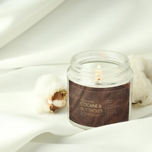 May include: A lit, clear glass jar candle with a brown label that reads "Cocaine & Buttholes." The candle sits on a white surface next to cotton bolls. The candle's flame is visible.