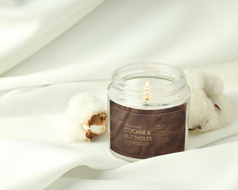 May include: A lit, clear glass jar candle with a brown label that reads "Cocaine & Buttholes." The candle sits on a white surface next to cotton bolls. The candle's flame is visible.