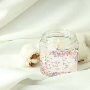 May include: A lit, clear glass jar candle with a floral label that reads "COCAINE & BUTTHOLES." The candle is surrounded by white fabric and cotton bolls. The label features pink, purple, and white roses.