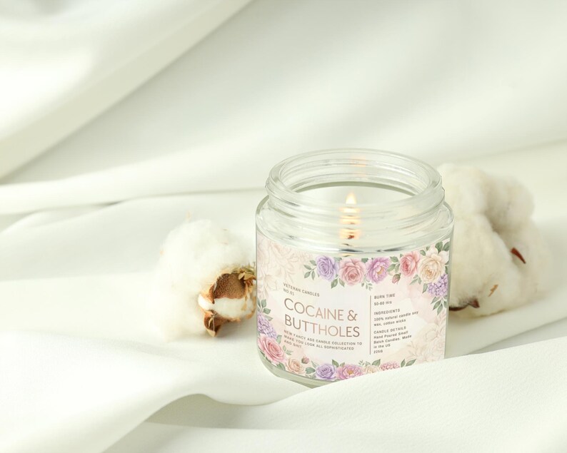May include: A lit, clear glass jar candle with a floral label that reads "COCAINE & BUTTHOLES." The candle is surrounded by white fabric and cotton bolls. The label features pink, purple, and white roses.