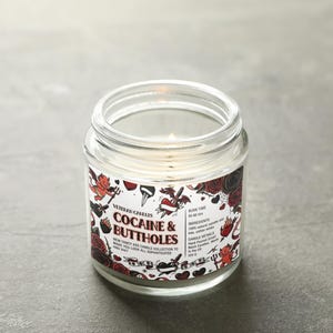 Może przedstawiać: Przezroczysta szklana świeca w słoiku z zapalonym knotem. Etykieta zawiera tekst "Cocaine & Buttholes" z ilustracjami w stylu tatuażu przedstawiającymi r&oacute;że, serca i diabły. Świeca stoi na szarej powierzchni.