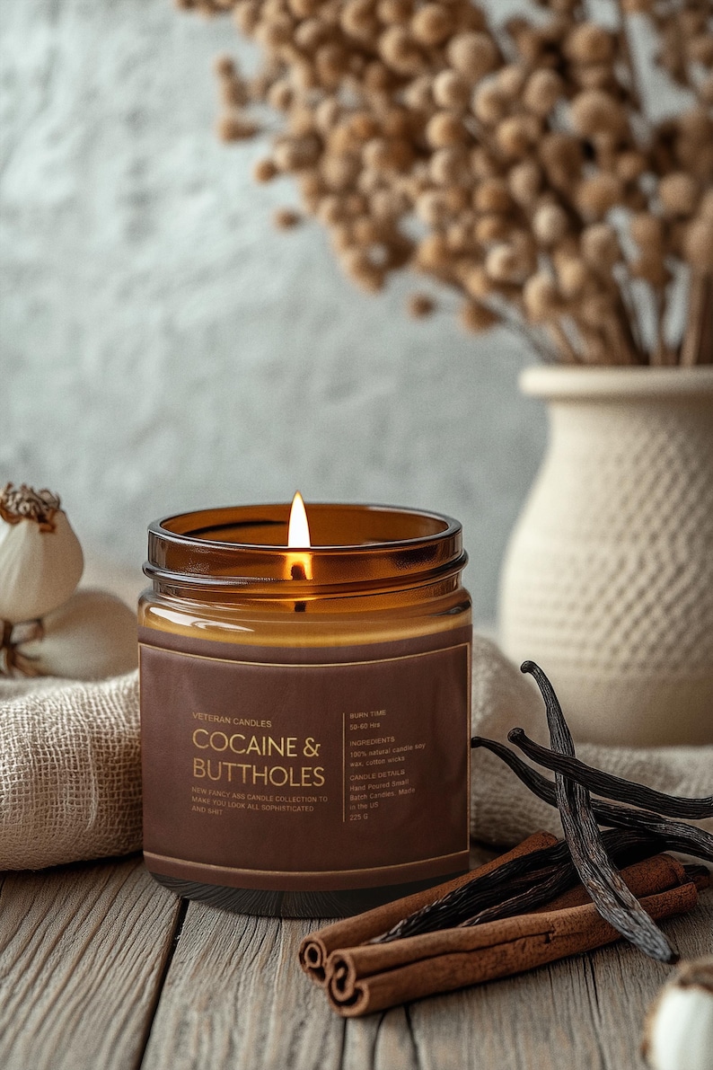 May include: A lit brown glass jar candle with the text "COCAINE & BUTTHOLES" on a brown label. The candle sits on a wooden surface with cinnamon sticks and vanilla beans. A vase with dried flowers is in the background.