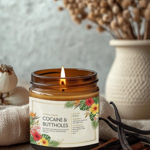 Może przedstawiać: Zapalone brązowe świeca w szklanym słoiku z etykietą z napisem "COCAINE & BUTTHOLES" z tropikalnymi wzorami kwiatowymi. Świeca stoi na drewnianej powierzchni z laskami cynamonu i strąkami wanilii. W tle wazon z suszonymi kwiatami.