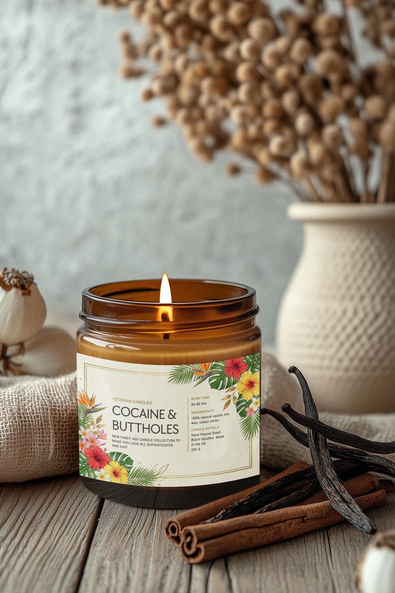 Może przedstawiać: Zapalone brązowe świeca w szklanym słoiku z etykietą z napisem "COCAINE & BUTTHOLES" z tropikalnymi wzorami kwiatowymi. Świeca stoi na drewnianej powierzchni z laskami cynamonu i strąkami wanilii. W tle wazon z suszonymi kwiatami.