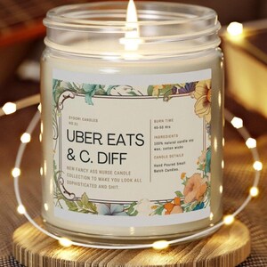 May include: A lit candle in a glass jar with a floral label. The label reads "UBER EATS & C. DIFF" and "New Fancy Ass Nurse Candle Collection." The candle sits on a wooden coaster, with string lights around it. The candle's burn time is 45-50 hours.
