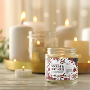 May include: A lit jar candle with the text "Cocaine & Buttholes" on the label, surrounded by roses, hearts, and cherubs. Several other lit candles of varying sizes are in the background, creating a warm, inviting ambiance.