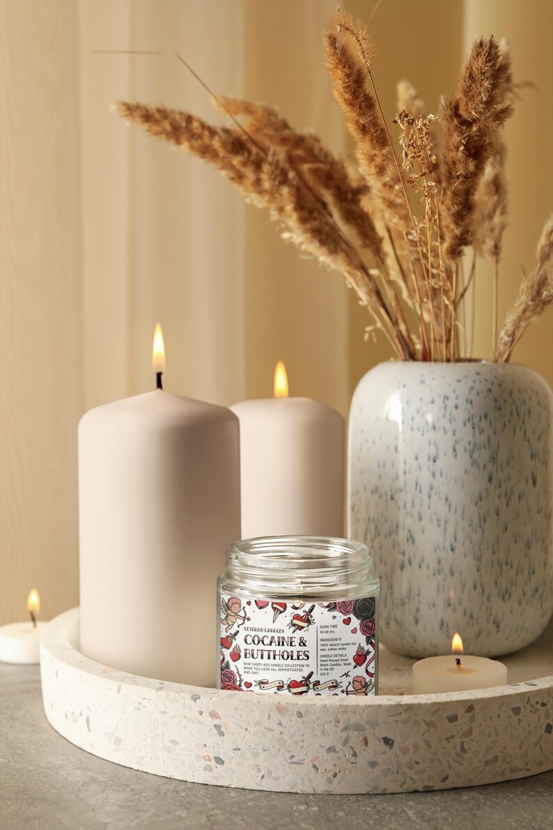 May include: A decorative arrangement featuring lit candles and a small glass jar candle with the text "COCAINE & BUTTHOLES". The candles are white and sit on a round tray. A vase with dried plants is in the background.