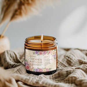May include: A lit candle in a brown glass jar with a floral label that reads "Cocaine & Buttholes." The candle sits on a tan cloth. The label has a floral design with pink, purple, and cream-colored roses.