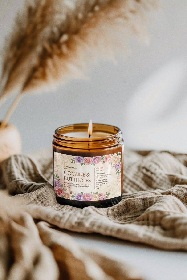 May include: A lit candle in a brown glass jar with a floral label that reads "Cocaine & Buttholes." The candle sits on a tan cloth. The label has a floral design with pink, purple, and cream-colored roses.