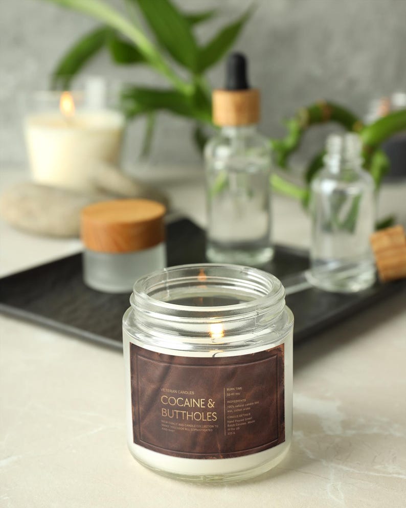 May include: A lit, clear glass jar candle with a brown label that reads "Cocaine & Buttholes". Other glass bottles and a wooden-topped jar are in the background. The candle is on a black tray.
