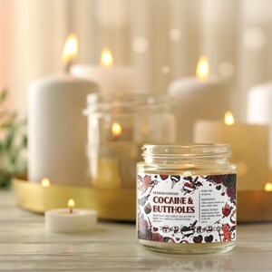 Może przedstawiać: Świeca w szklanym słoiku z etykietą z napisem "Cocaine & Buttholes" z dekoracyjnym wzorem. Świeca otoczona jest zapalonymi świecami o r&oacute;żnych rozmiarach i złotą tacą. Tło jest rozmyte.
